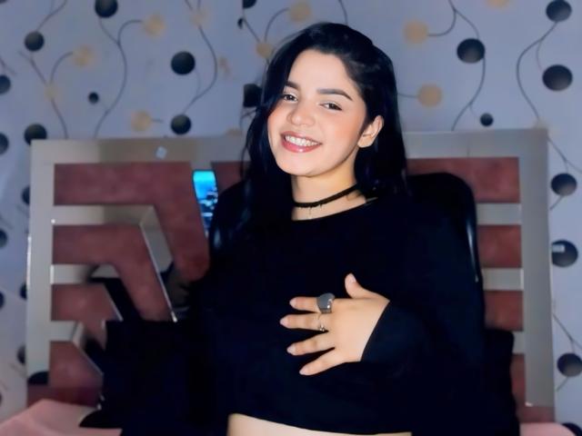 EvaGoldX with black hair, smiling, wearing a black long-sleeve top, choker, and ring, covering her tits with one hand, standing in front of a pink and white bed.