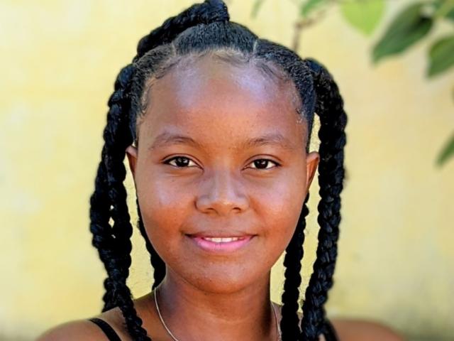 Eliani has dark brown skin, black braided hair in two long twists, and a small smile with pink lipstick. She's wearing a black top and a silver necklace.