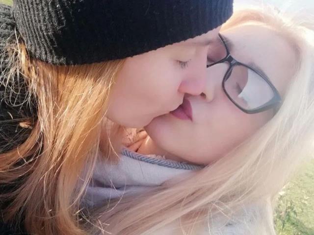 DoubleloveXXX-hot with blonde hair and glasses kisses DoubleloveXXX-hot with brown hair and a black beanie. Their noses touch, eyes closed. She's wearing a gray hoodie.