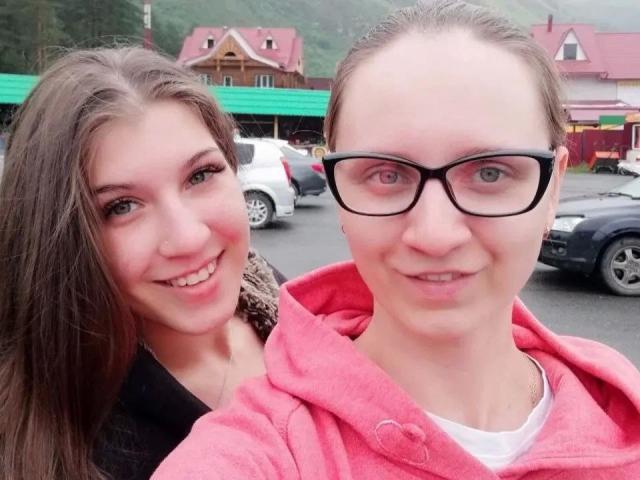 DoubleloveXXX-hot with brown hair and DoubleloveXXX-hot with glasses and pink hoodie, both smiling. Boobs are not visible, focus is on their faces. Background includes parked cars.