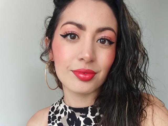 ValeryX69 has pink eye makeup, red lipstick, and black eyeliner with thick lashes. She's wearing a leopard print top and large gold hoop earrings, with wavy dark hair.