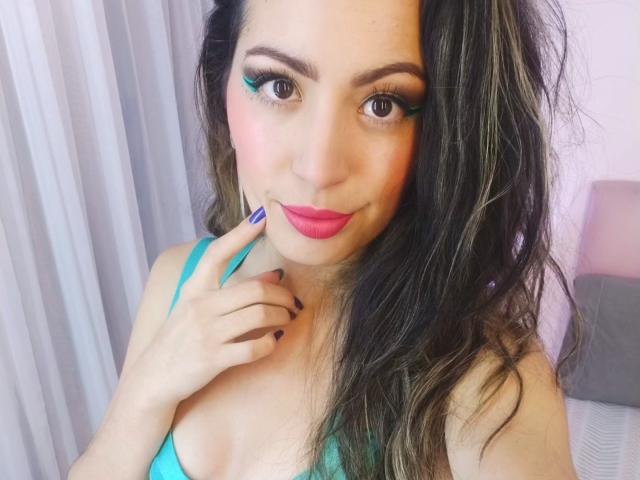 ValeryX69 with pink lipstick, blue eye makeup, long wavy brown hair, turquoise bra, holding finger to cheek, purple nails, smiling at camera. Boobs slightly visible.