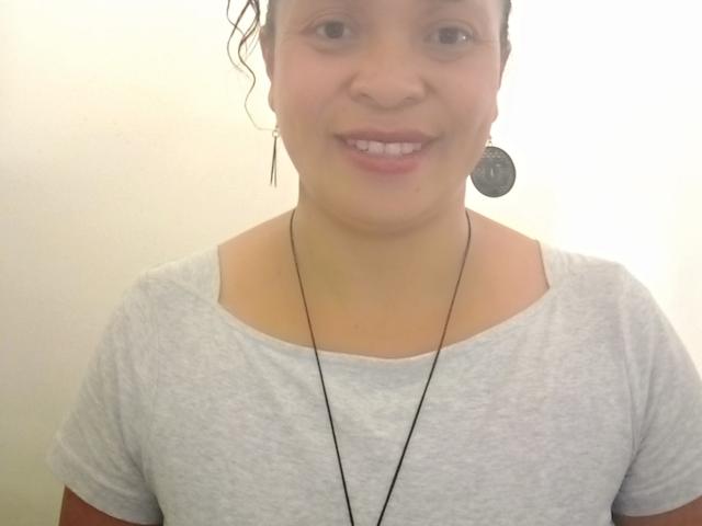 Rahel69 with curly hair, light skin, small nose, gray shirt, black necklace, smiling, wearing round earrings, white background.