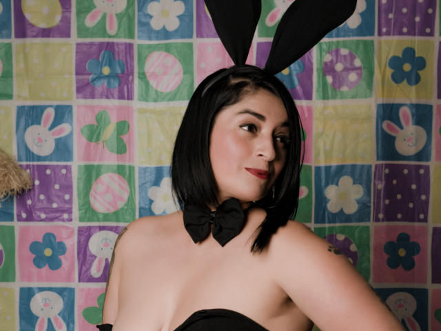 KarenBroc with black bunny ears, short black bob, red lipstick, black strapless top, bow tie, and a slight smile, showing her ass and tits. Her skin is pale.