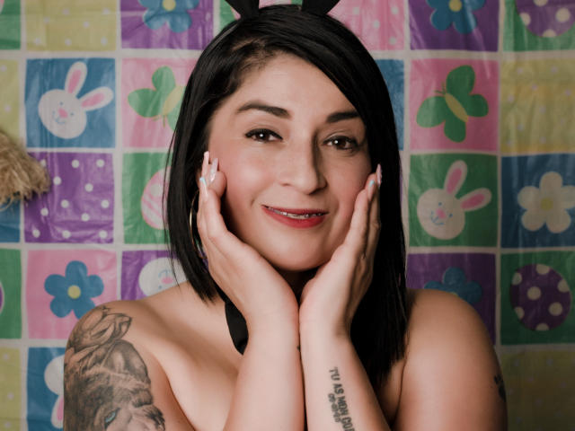 KarenBroc has short black hair, bunny ears, tattoos, and is smiling with hands on her face, showing off her small tits and pierced lip. She's topless with pink lipstick.