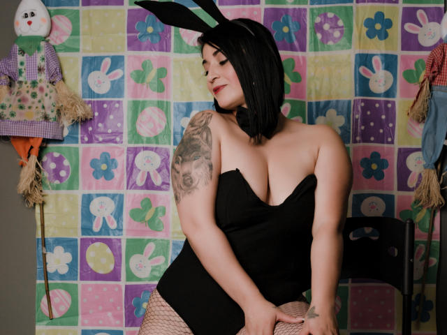KarenBroc with black bob, wearing bunny ears, black strapless top, fishnet stockings, showing tattooed arm, and smiling, with hands on knees, showing her tits.