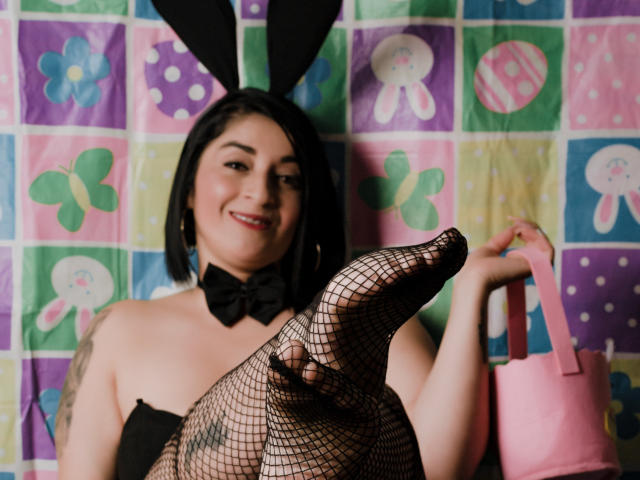 KarenBroc with black bunny ears, fishnet stockings, and a pink bag, smiling at the camera, showing her foot and wearing a black bowtie, has visible tattoos.