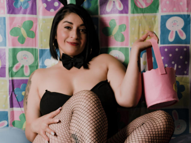 KarenBroc with black bob, bowtie, black strapless top, fishnet pants, holding pink purse, sitting, smiling, tattoo on left arm, short black hair, pink background.
