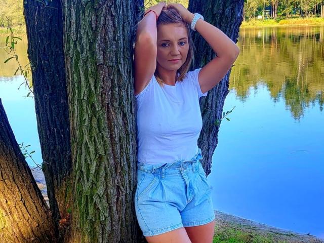 SexiSeal leans against a tree with arms raised, wearing a white t-shirt and blue shorts, standing by a reflective blue lake, with a white bracelet on her wrist.