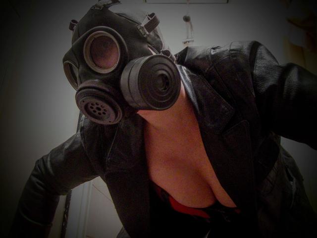 MistressOnline-hot in black leather jacket, red bra, and green gas mask, leaning forward, showing her tits, with her face hidden by the mask, in dim lighting.
