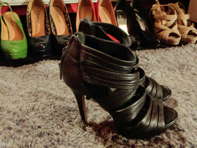 Black leather high-heeled shoe with multiple straps and a zipper, standing on a gray carpet, with various colorful and patterned heels in the background. MistressOnline-hot's shoe.