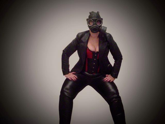 MistressOnline-hot in black leather suit, red corset, and gas mask, standing with legs apart, hands on hips, showing small tits, black leather pants, and confident stance.