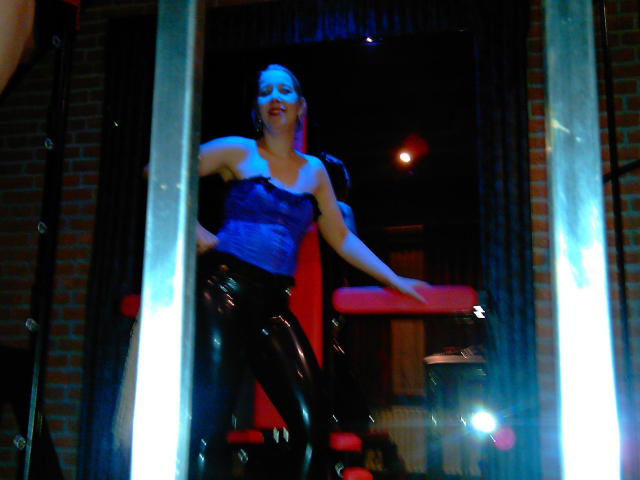 MistressOnline-hot with blue face paint, purple top, and shiny black latex pants, dancing in a dimly lit room with blue and red lights, flanked by metal pillars.