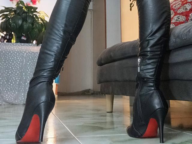 CharlizeLegs is wearing black thigh-high leather boots with red soles, standing on a tiled floor. Boots are zipped, showing her legs and high heels.