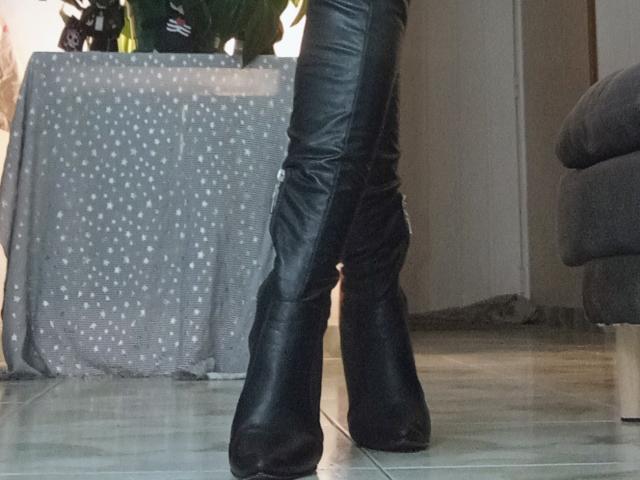 CharlizeLegs' black leather thigh-high boots with chunky heels, standing on a tiled floor, showing the back of her legs and boots. Boobs not visible, ass not visible.