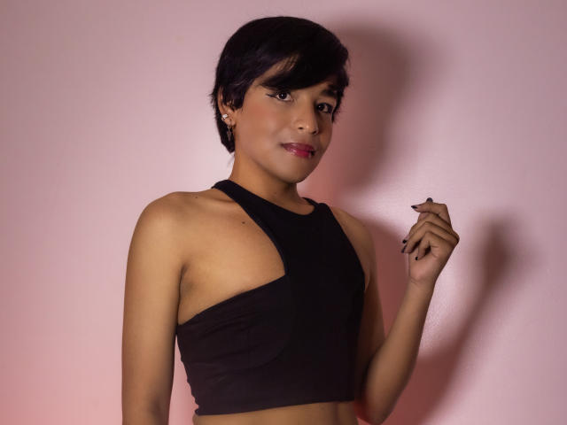 DannyHotz with short black hair, wearing a black halter top, showing bare shoulders, and holding a nail with black nail polish, smiling with pink lipstick.