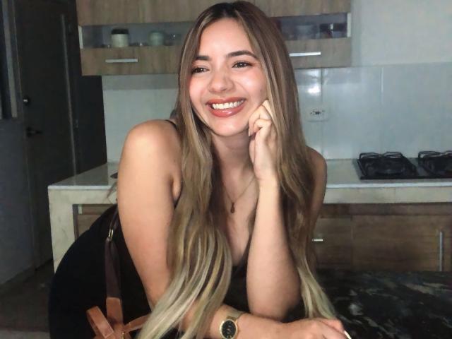 MajoGomez, con lunghi capelli biondi, sorridente, appoggiata a un bancone della cucina, indossa un vestito nero, una borsa a tracolla marrone e un orologio, mostrando il seno e i glutei..