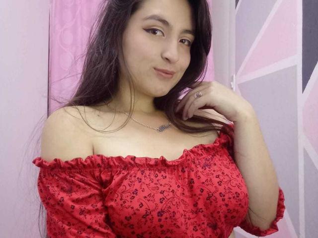 VenusSweetPalmer with long brown hair, red off-shoulder dress, small black floral print, ring on right hand, subtle smile, slight pout, necklace, and medium-sized tits.