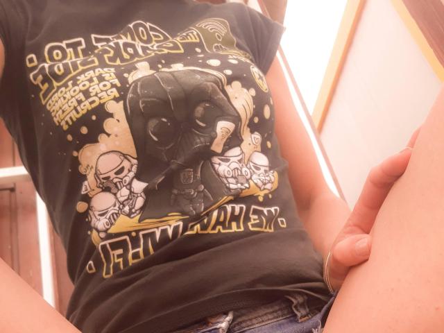 Aiyana's black t-shirt with a Darth Vader and Stormtrooper graphic, showing her small, erect, uncircumcised penis, sitting with legs apart, wearing blue jeans, and a gold bracelet.