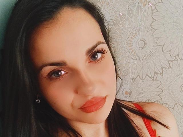 O rosto de MelissaXHotty, com cabelo castanho escuro, olhos azuis e batom laranja, vestindo uma blusa vermelha, olhando para a câmera com um leve sorriso, mostrando seus seios..