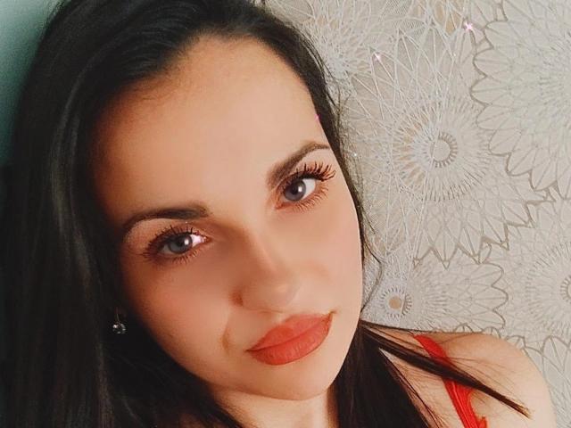 Le visage de MelissaXHotty, avec ses cheveux bruns foncés, ses yeux bleus et son rouge à lèvres rouge, mettant en valeur ses cils et ses lèvres légèrement entrouvertes, portant un haut rouge, regardant l'appareil photo..