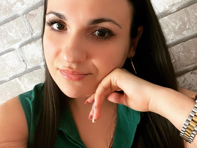 MelissaXHotty, com cabelos longos e escuros, camisa verde e pulseira de prata, apoiando o queixo na mão, olhos castanhos, sorriso discreto e batom rosa, usando brincos de argola dourados..