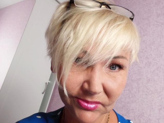 HellyHots-hot has short, blonde hair with a side part, pink lipstick, blue eyes, and a blue shirt with white polka dots. Eyeglasses rest on her head.