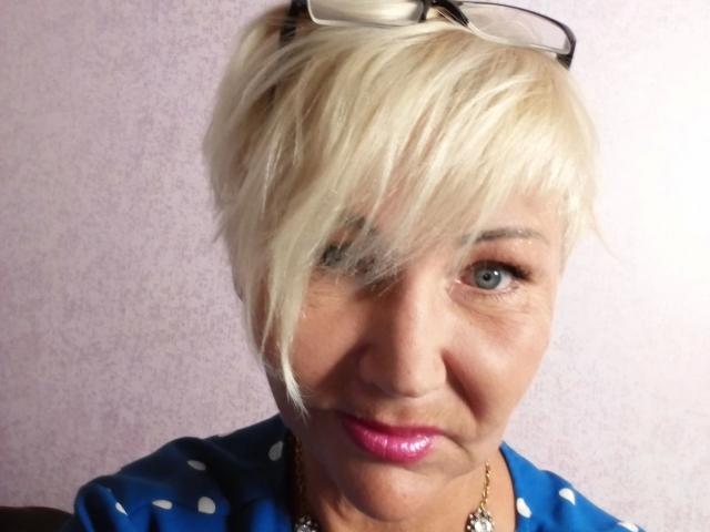HellyHots-hot has short blonde hair, blue eyes, pink lipstick, and wears a blue polka-dot shirt with black glasses on her head and a gold necklace.