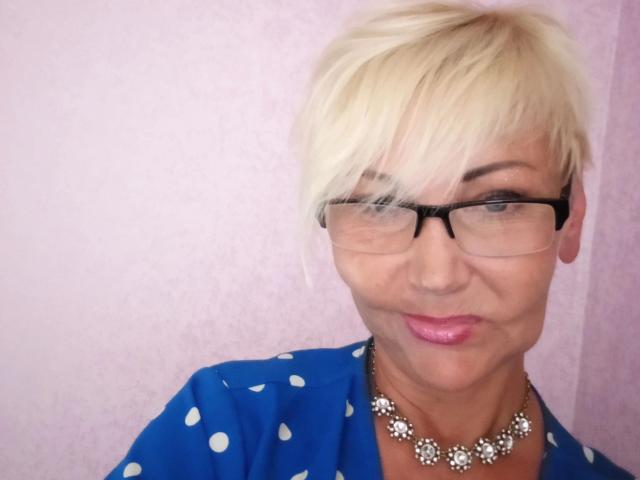Blonde HellyHots-hot with short hair, black glasses, pink lipstick, blue polka dot shirt, and flower necklace, giving a slight smile. Her ass is not visible.