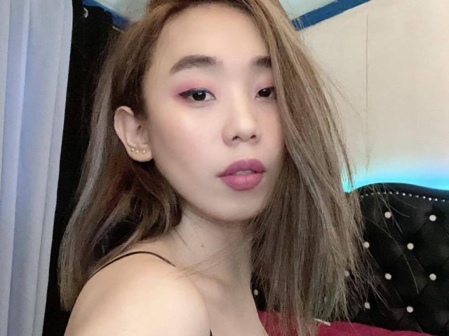 MissSultryAya, met roze oogschaduw, roze lippenstift en licht open lippen, heeft steil bruin haar, kleine oorbellen en een zwarte band, en kijkt met een neutrale uitdrukking naar de camera..