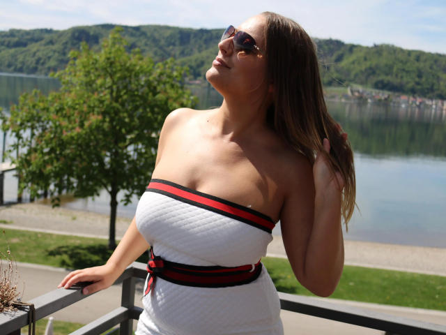 Lollipop69-hot with tanned skin, medium boobs, and long brown hair in a white strapless dress with red and black stripes, wearing sunglasses, standing by a railing, holding hair.