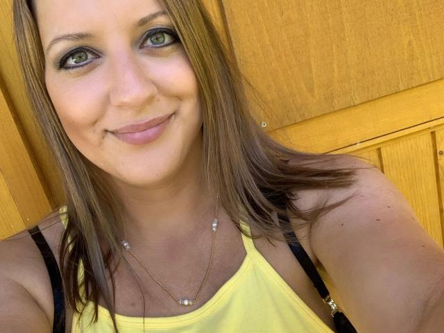 Lollipop69-hot with brown hair, green eyes, and a yellow tank top, smiling at the camera, showing her tits slightly in the black straps, wearing a delicate necklace.