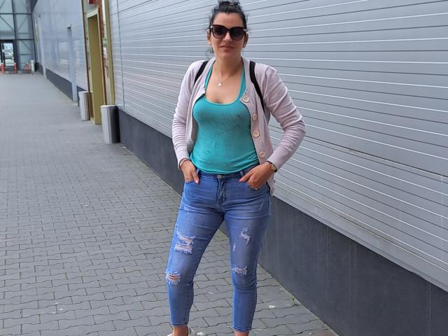 KissYouKissMe has tits, wearing a teal tank top, ripped blue jeans, pink cardigan, and sunglasses, standing with hands in pockets on a gray brick sidewalk.