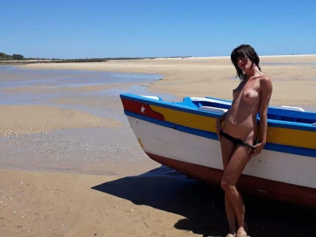 Nude Aiyana with small tits and black panties stands by a colorful boat on a sandy beach, her right hand on her hip, under a clear blue sky.