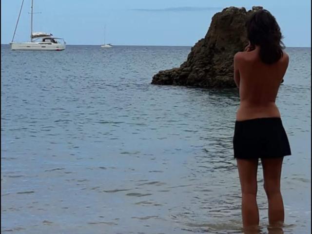 Topless Aiyana with black skirt stands in shallow water, back to camera, facing rocky outcrop and two boats on calm sea, under blue sky. Aiyana has brown hair.