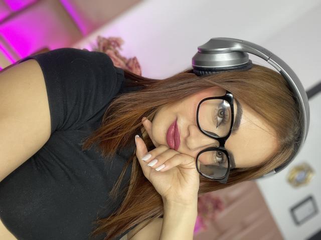 AngelCutie with glasses, brown hair, pink lips, black t-shirt, gray headphones, resting chin on hand, looking at camera, white nails, pink highlights in background.