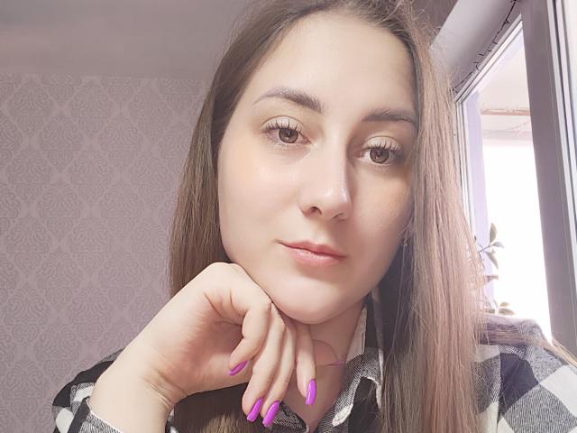NikaCocpit has light skin, brown eyes, and long brown hair. She's wearing a plaid shirt, with purple nail polish, resting her chin on her hand, looking at the camera.