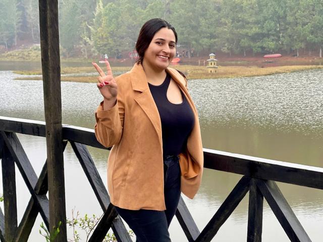 KattaAndJhon has medium tits, black tank top, tan coat, blue jeans, and dark hair, smiling, holding up a peace sign, standing by a wooden bridge by a lake.