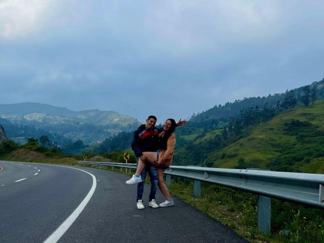 Katta e Jhon sono in piedi su una strada, Katta con i glutei in evidenza, Jhon la tiene, entrambi sorridono. Katta indossa un vestito color sabbia, Jhon una giacca blu. Il cielo è nuvoloso, sullo sfondo si vedono le montagne..