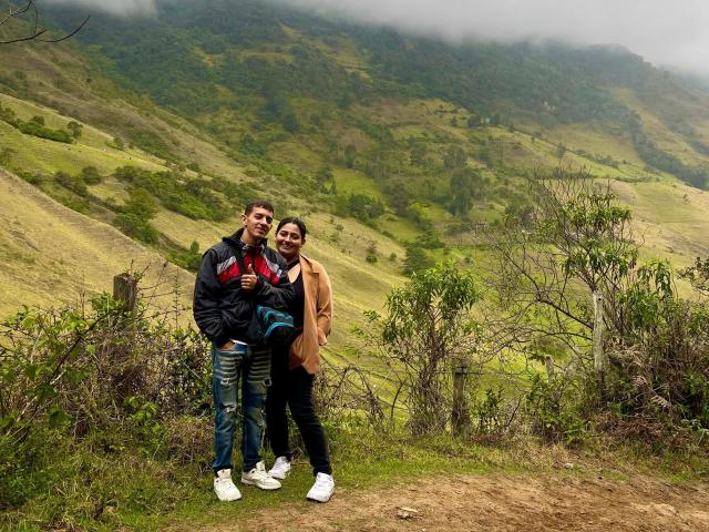 KattaAndJhon, de pie en un sendero de tierra, vistiendo jeans y chaquetas, uno con un abrigo marrón, sonriendo, sosteniendo una cerveza, fondo de montañas brumosas, colinas verdes, cielo nublado..