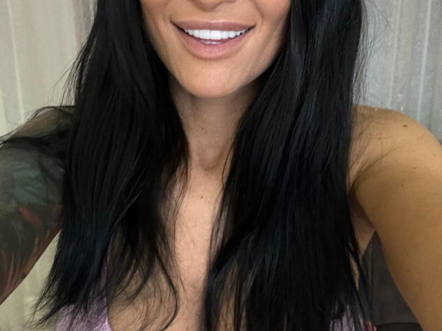 AndraD has long black hair, white teeth, and a tattoo on her right arm. She's smiling with visible cleavage in a pink top.