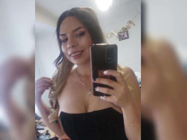 VioletaStone mit welligem, blondem Haar, schwarzem Top ohne Träger, lächelt in ein Spiegel-Selfie, hält ein Handy, freizügige Kleidung, mittelgroße Brüste, helle Haut, dezentes Make-up, Halskette, Schlafzimmer, unscharfer Hintergrund..
