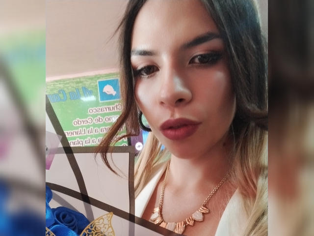 VioletaStone has brown eyes, full pink lips, and long brown hair with blonde ombre. She's wearing a gold necklace and a white top, looking slightly down.