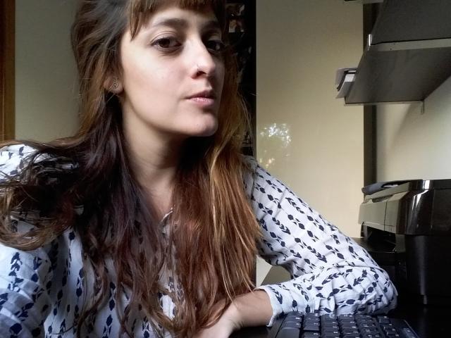 JaneRoa, avec de longs cheveux bruns, portant une chemise blanche à motifs, assise à un bureau, regardant légèrement vers la gauche, avec une imprimante et un clavier en arrière-plan..