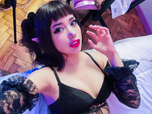 SabriPrettty with dark hair in pigtails, black lace bra, and arm sleeves, showing off her tits and pink lipstick, holding her hand up. Wooden floor, purple chair in background.