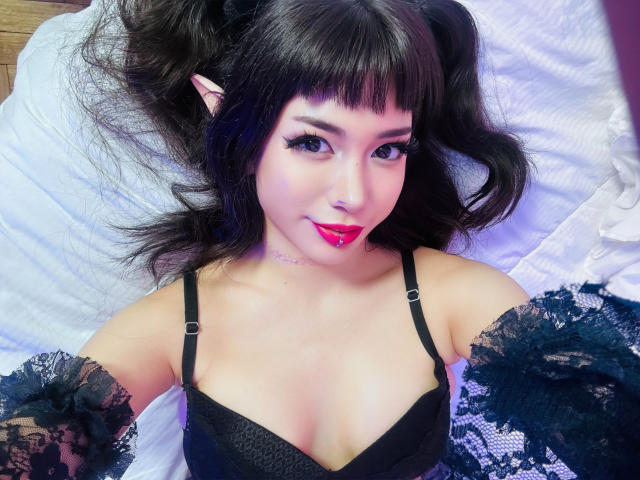 SabriPrettty with black lace sleeves, black bra, pink lipstick, and dark pigtails, lying on a white bed. Her pointy elf ears and cute face are visible. Boobs.