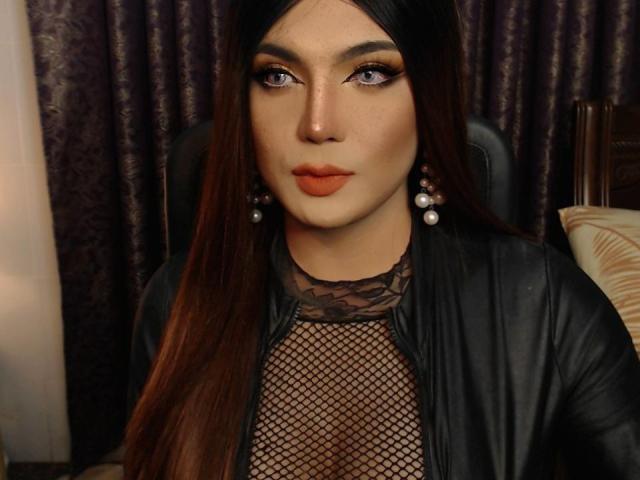 PattyHugeCock with long brown hair, orange lipstick, and fishnet top over black leather jacket, wearing pearl earrings, looking at camera with serious expression.
