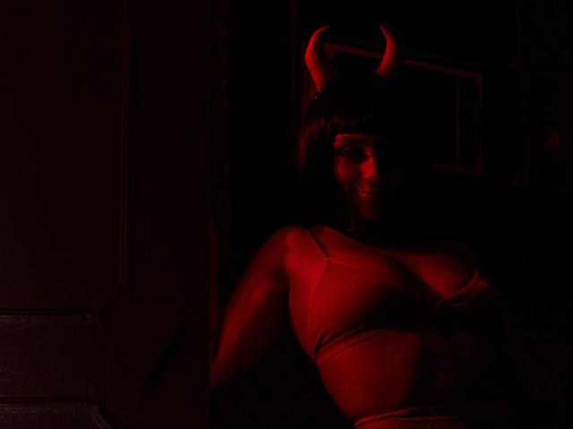 Zdiablaz with red horns, wearing a tight, red, strapless top, sitting in dark, red-lit room, smiling, showing small, perky tits, and a cute, sexy face.
