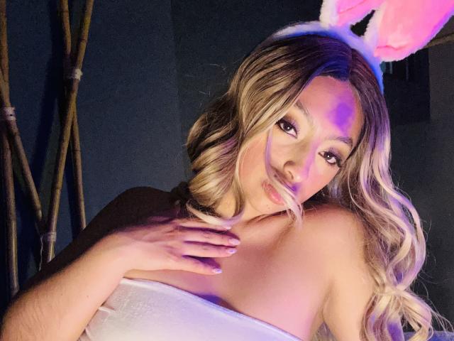 Zdiablaz with blonde wavy hair, pink bunny ears, and a white strapless top, pouting with a tongue out, hand on chest, purple and blue light on face.