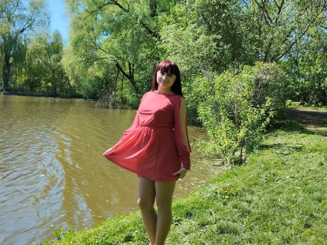 NinaFlawlees stands by a river, wearing a red dress, holding it out, showing her bare ass and pussy. She has long brown hair and green eyes.
